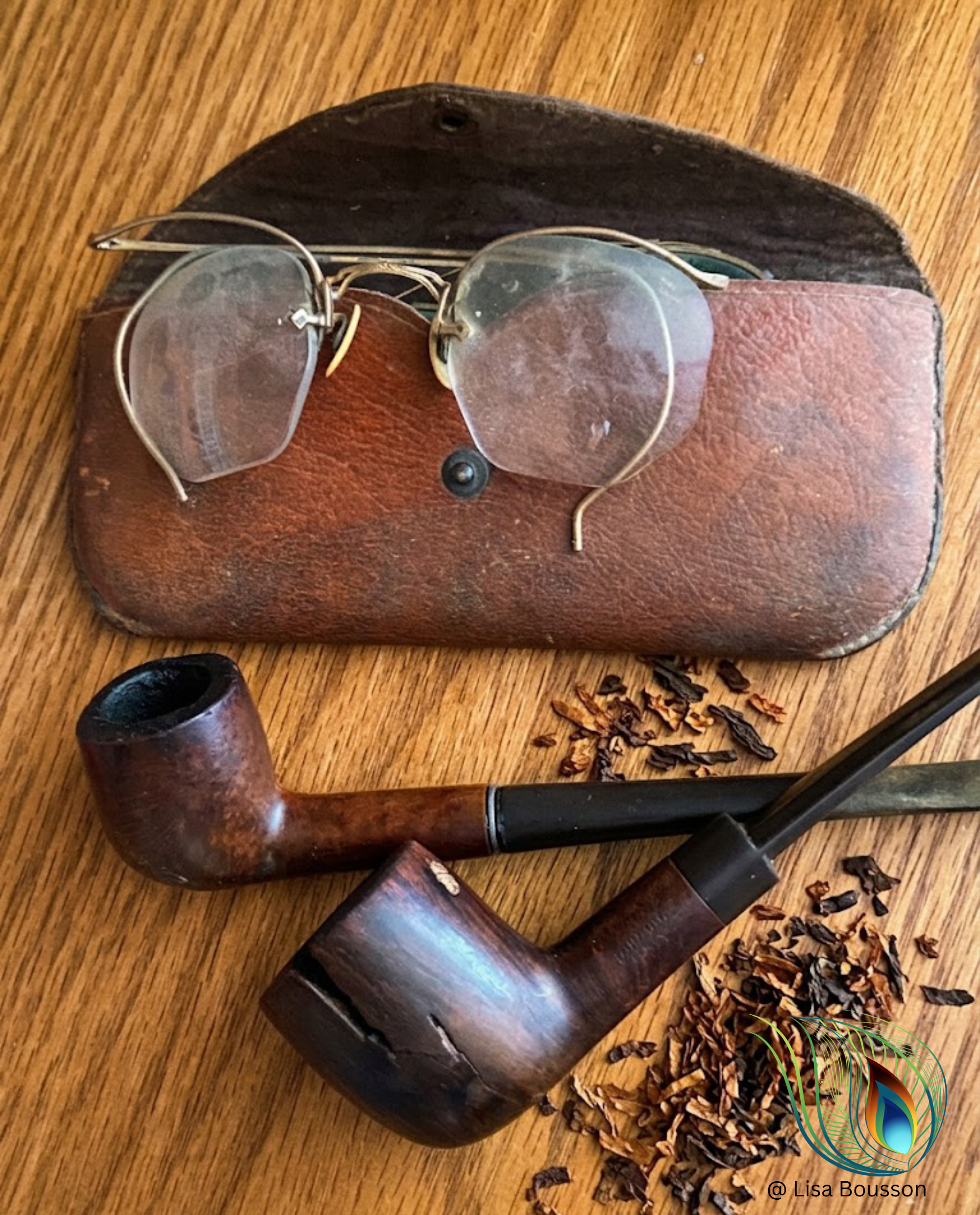 An antique pair of round glasses on a antique glasses case. Nearby are two antique pipes with loose tobacco nearby.